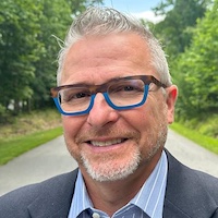 a man with grey facial hair, grey hair glasses wearing a suit standing the the middle of a road. Headshot. 