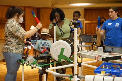 Music students encourage a 5-year-old camper while his mother and younger brother look on.