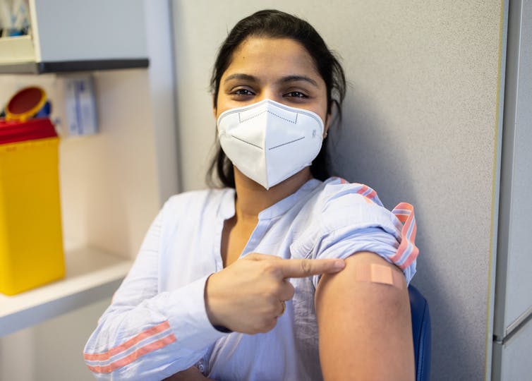 Woman smiles and points at band aid on arm where she just received a vaccination