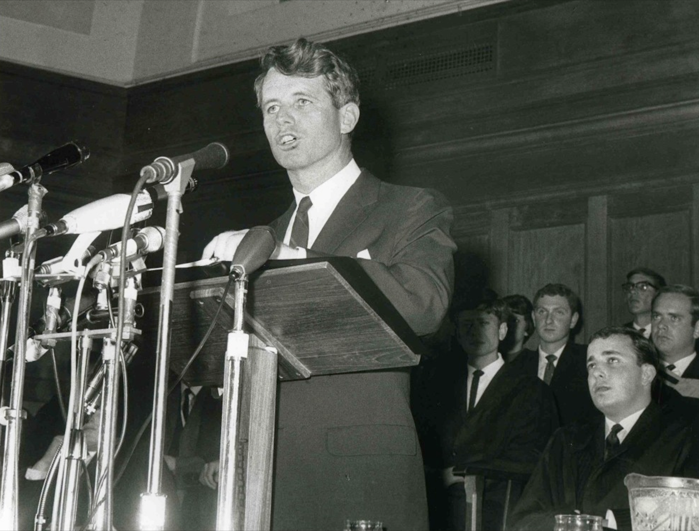 RFK wears a suit delivering a speech into multiple microphones.