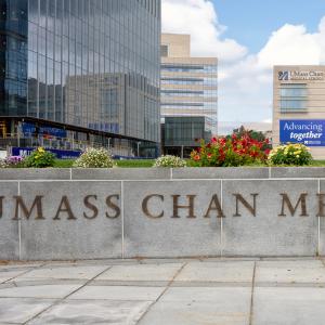 UMass Chan Medical School sign on a curved wall with the campus in the background on a warm summer day. 