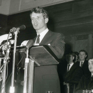 RFK wears a suit delivering a speech into multiple microphones.