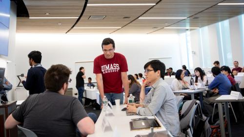 Classroom filled with people as a person wearing a UMass Amherst shirt talks with two people seated at a table.