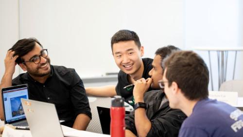 Four students sitting at a desk, talking together with an open laptop.