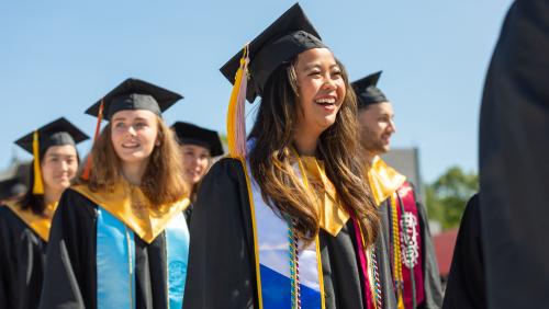 Students wearing graduation regalia stand together outdoors, smiling.