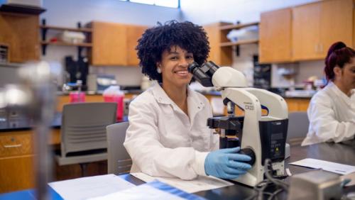 Student in a lab smiling at the camera while using a microscope.